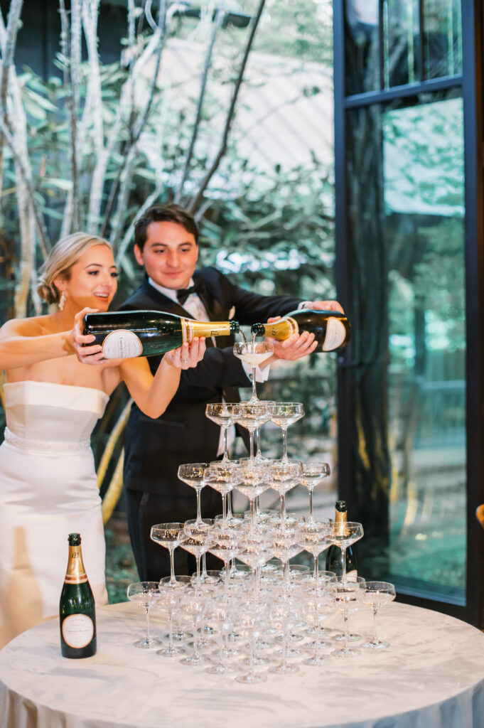 A bride and grooming pouring champagne into stacked coupe glasses at a wedding reception at The Farm at Old Edwards Inn in Highlands, North Carolina. 