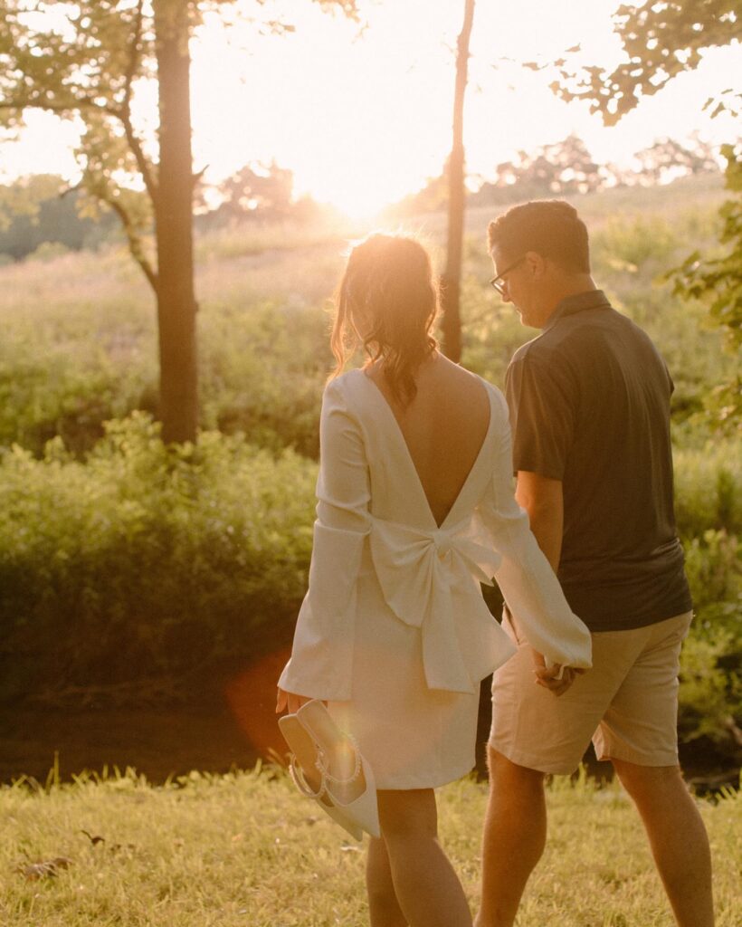 A woman in a white dress carrying her shoes and holding hands with a man. Both are walking away from the camera beside a creek in Asheville, North Carolina. 