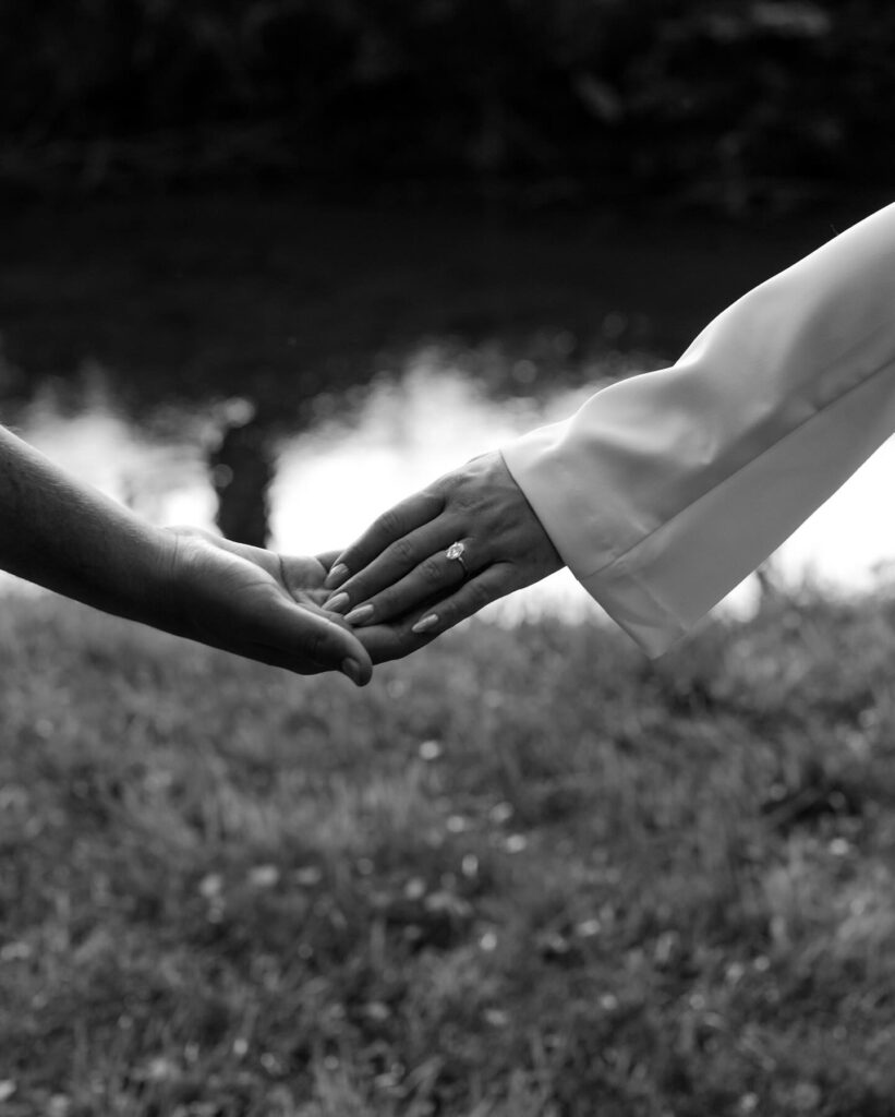 A woman's hand with an engagement ring, holding a man's hand next to a creek in Asheville, North Carolina. 