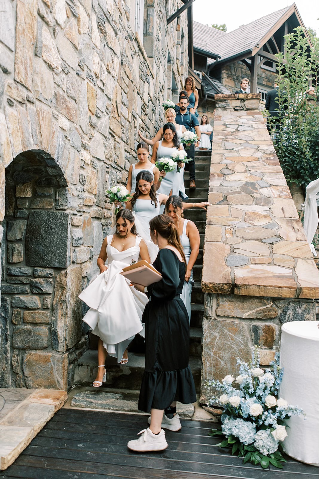 A wedding planner dressed in black standing at the bottom of an outdoor stone staircase as a bride and her bridesmaids walk down them. 