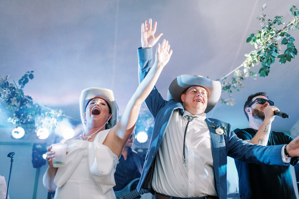 A bride and groom, both wearing white cowboy hats, dance on  a stage with their arms raised at Trillium Links and Lake Club in Cashiers, North Carolina.