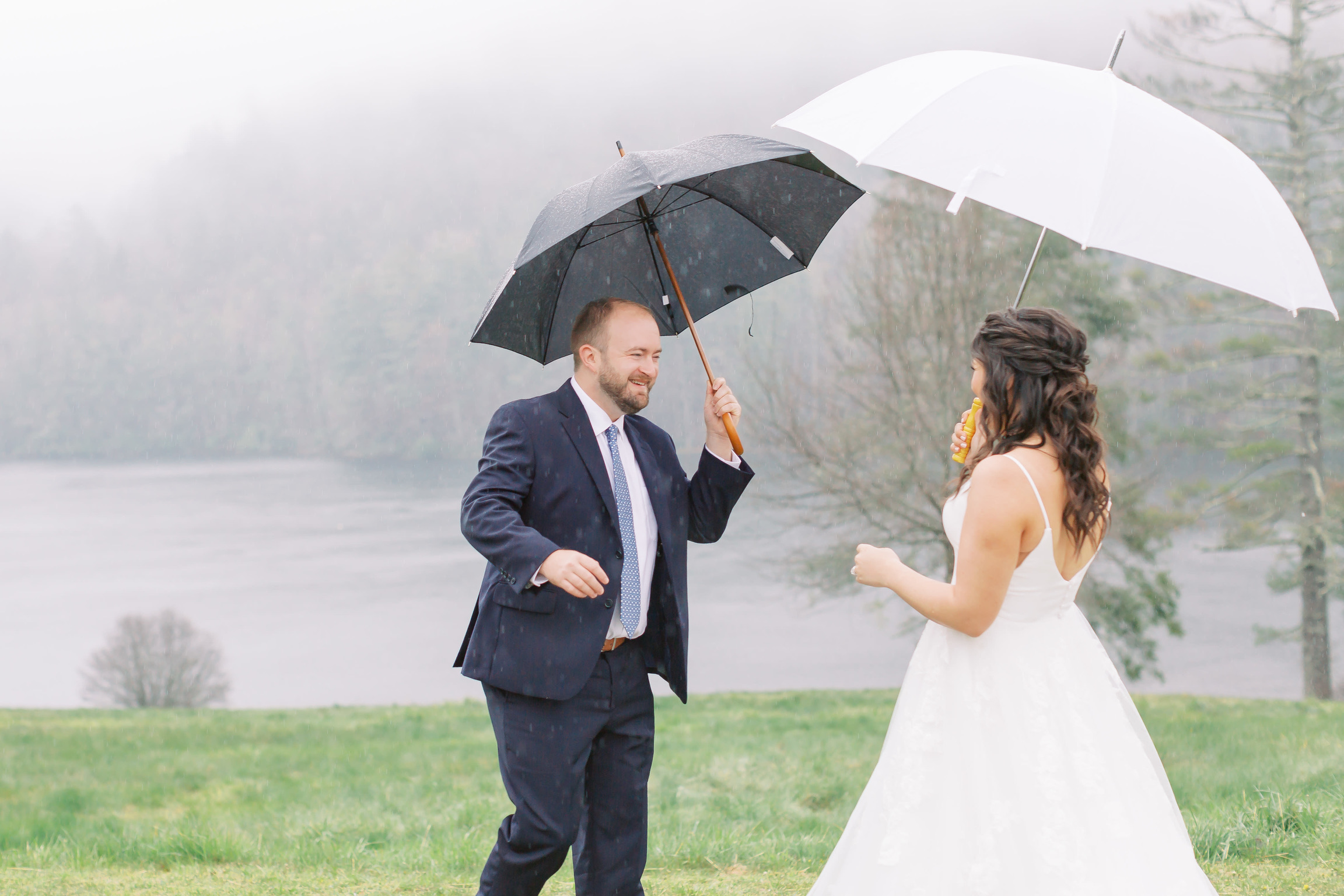 Bride and groom laughing under umbrellas on a misty day at a lakeside mountain wedding in Western North Carolina